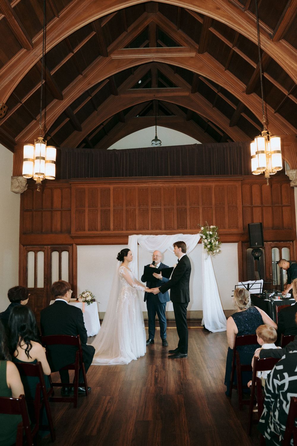 Wide view of wedding ceremony in grand hall with exposed wooden trusses and wall paneling.