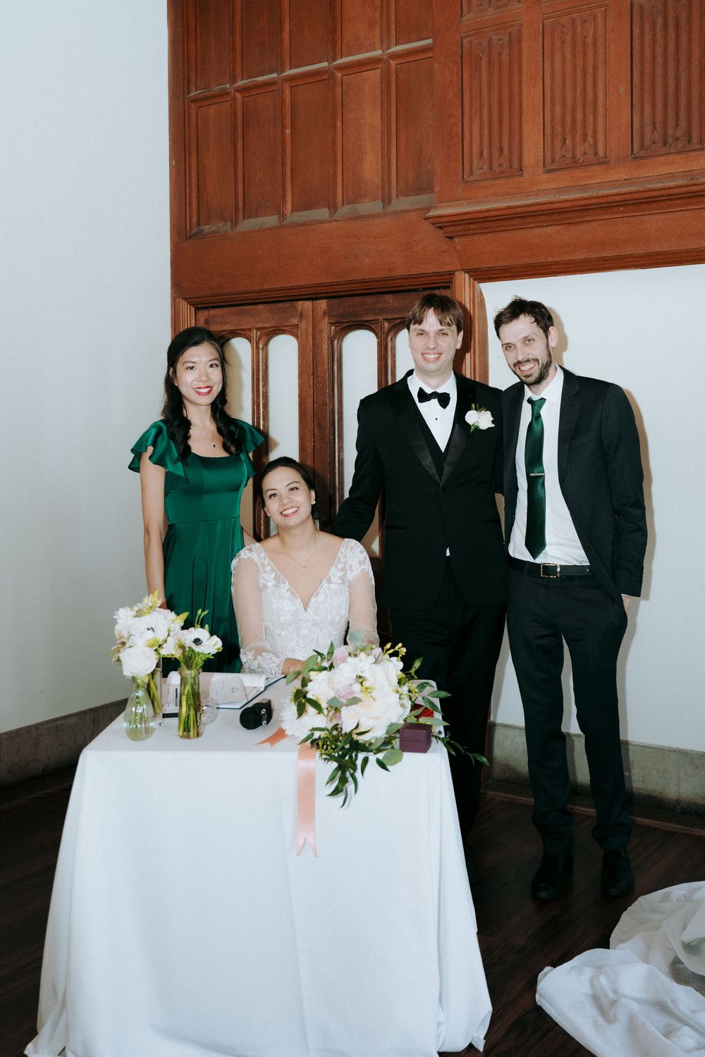 Wedding party gathered around white-clothed table with floral arrangements and emerald green accents.