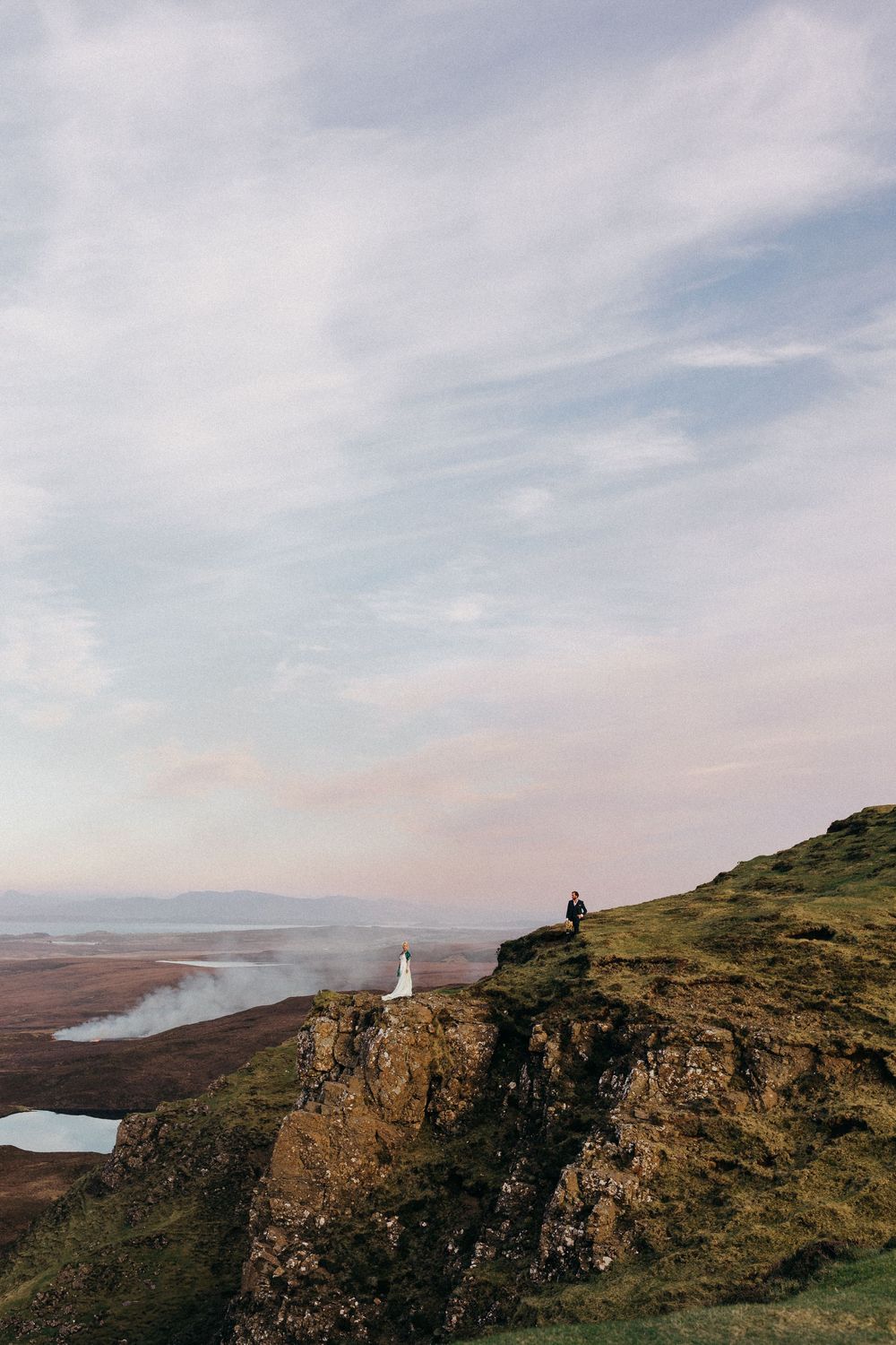 A lone figure sits atop dramatic coastal cliffs overlooking a misty valley with mountains in the distance under cloudy skies.