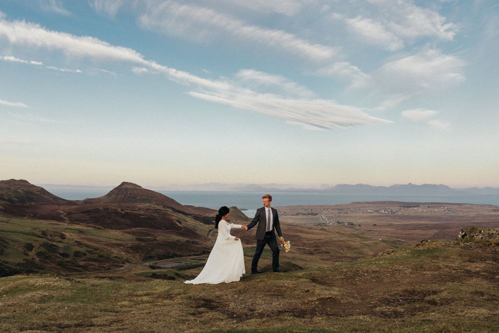 A couple stands on a hillside overlooking a scenic mountain landscape during golden hour under a cloudy sky.