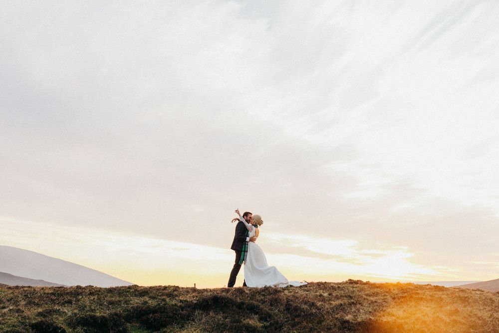 A couple embraces on a hilltop silhouetted against a glowing sunset with mountains in the distance.