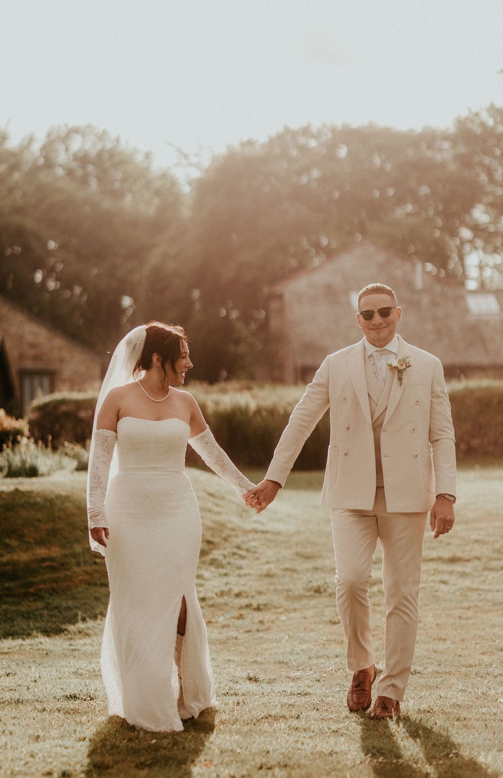A bride and groom walking hand in hand at sunset in a romantic outdoor garden setting.