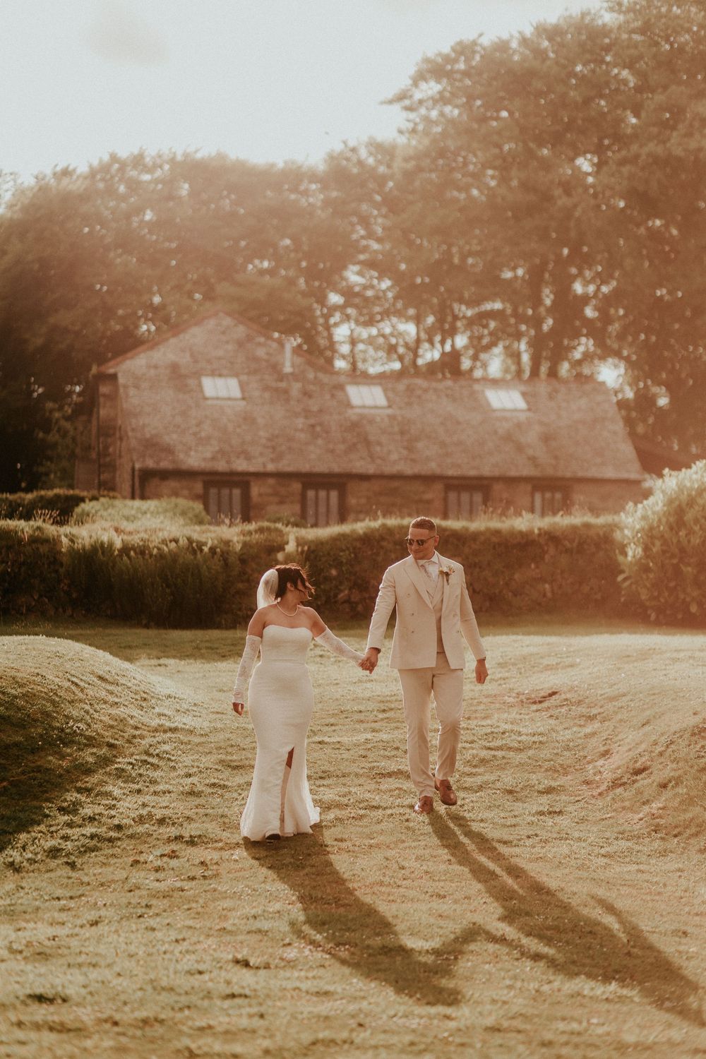 Two newlyweds walking hand in hand through a sunny field with a rustic cottage in the background.