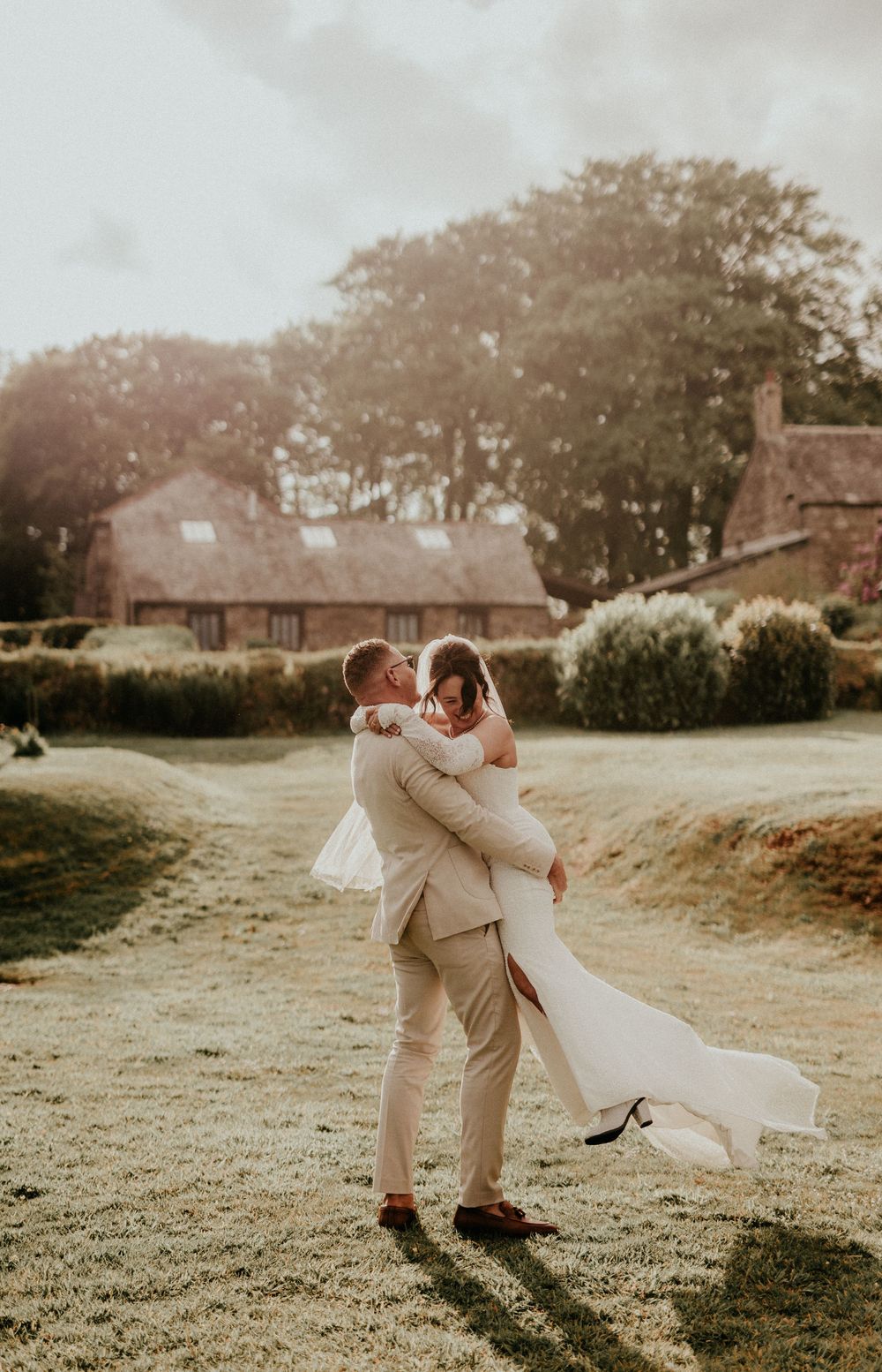 A romantic wedding couple embraces in a sunlit garden with historic buildings in the background at sunset.