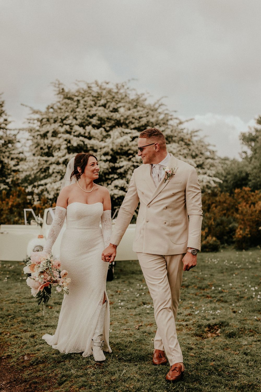 Couple in light colored wedding attire walking hand in hand on grassy field with blooming trees in background during sunset.