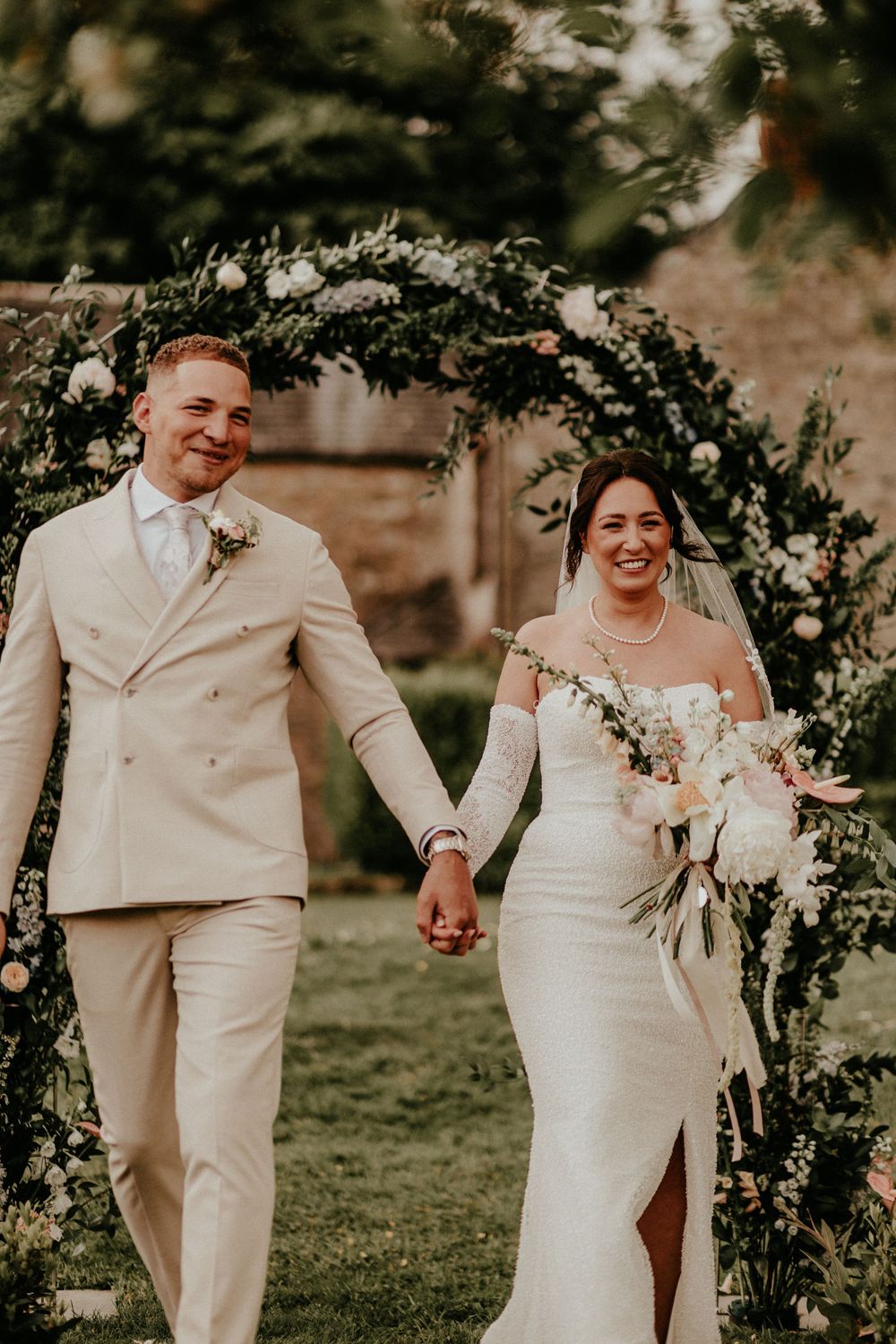 A joyful couple in wedding attire walking beneath a floral arch decorated with white flowers in a garden setting.