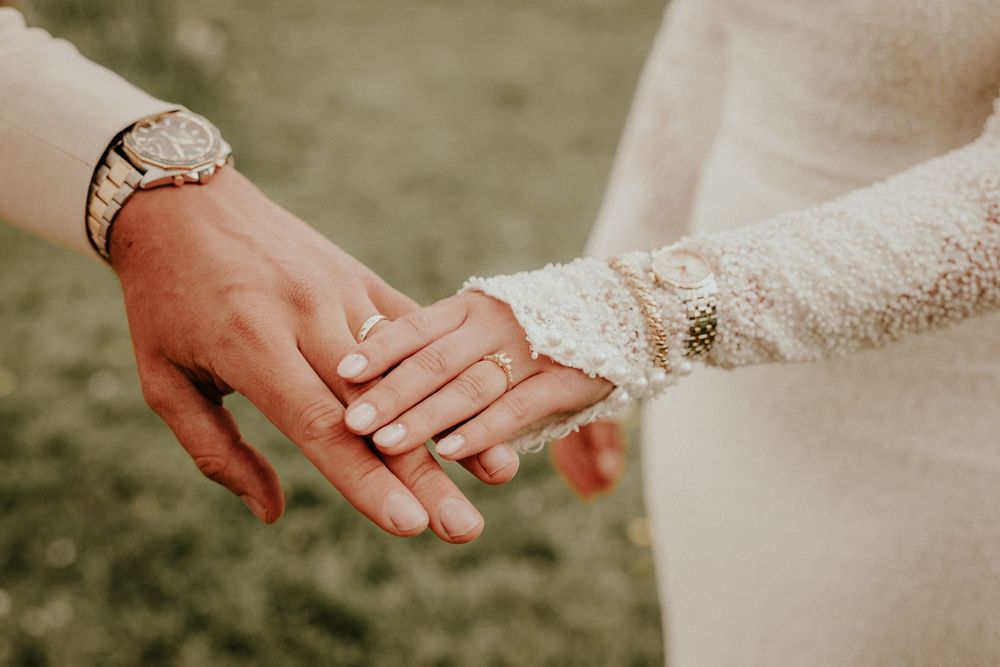Two hands wearing wedding rings touch tenderly against a soft natural background with lace sleeve detail.