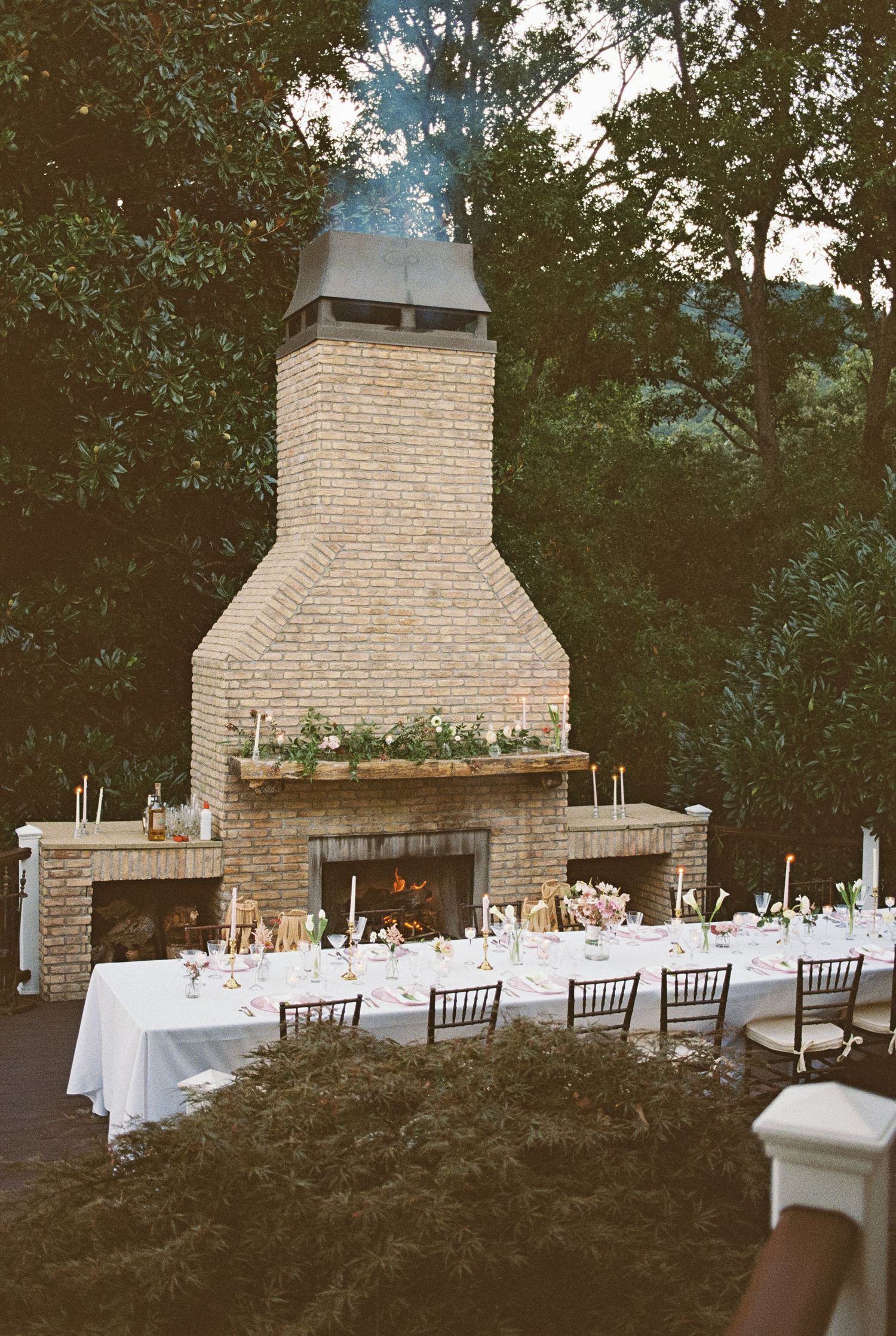 An elegant outdoor wedding reception table set against a large brick fireplace with white linens and place settings.