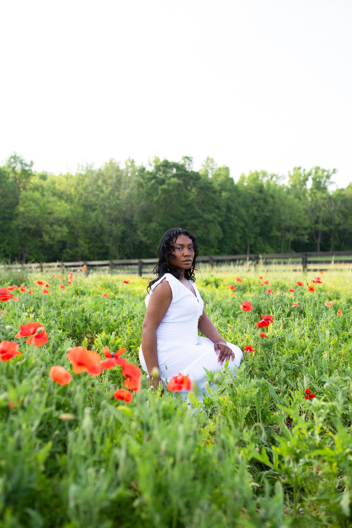 A woman in a white dress poses in a vibrant field of red poppies and green foliage on a sunny summer day.