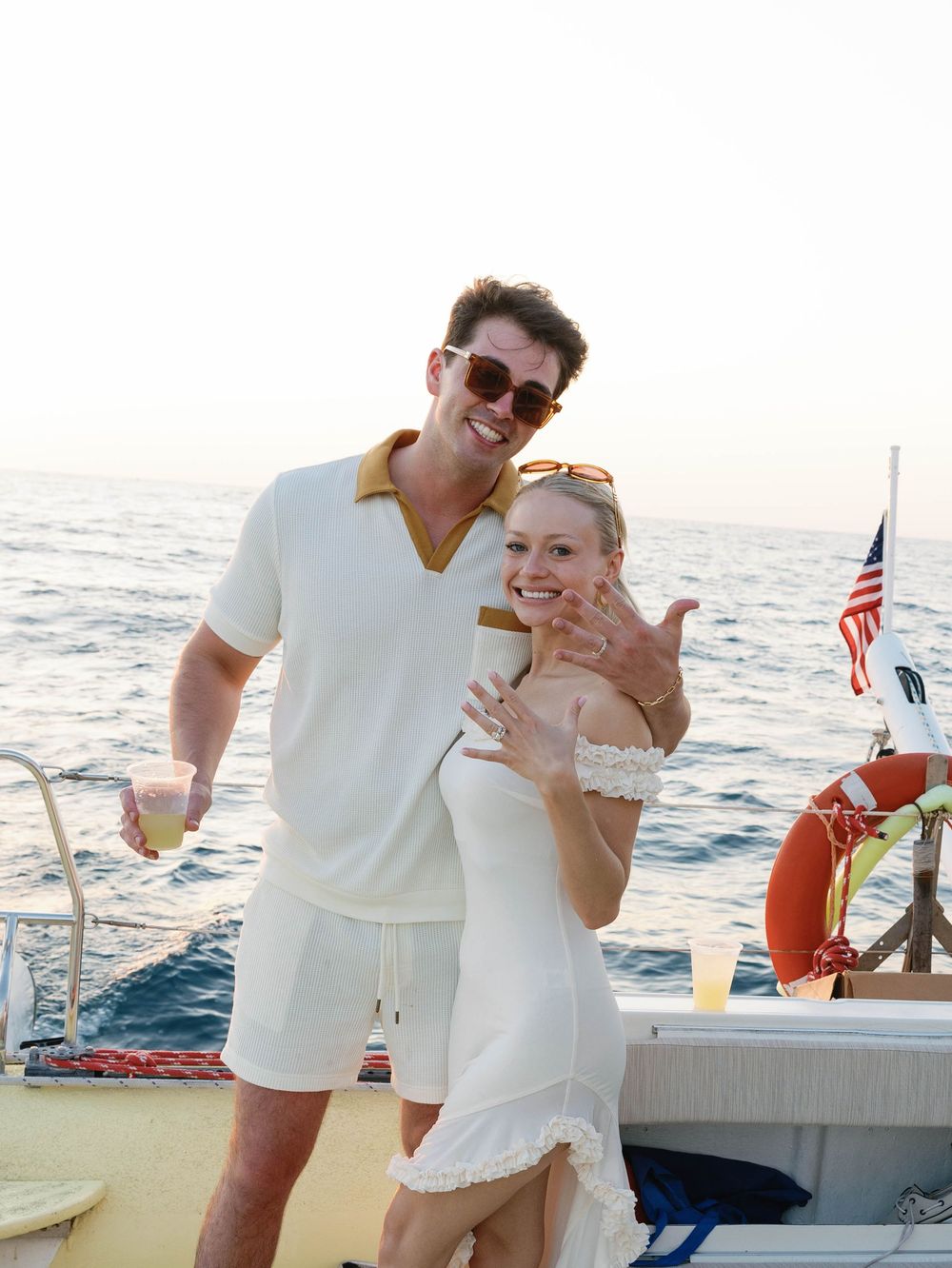 A couple in summer white outfits enjoying a sunny day on a yacht deck with an American flag visible.