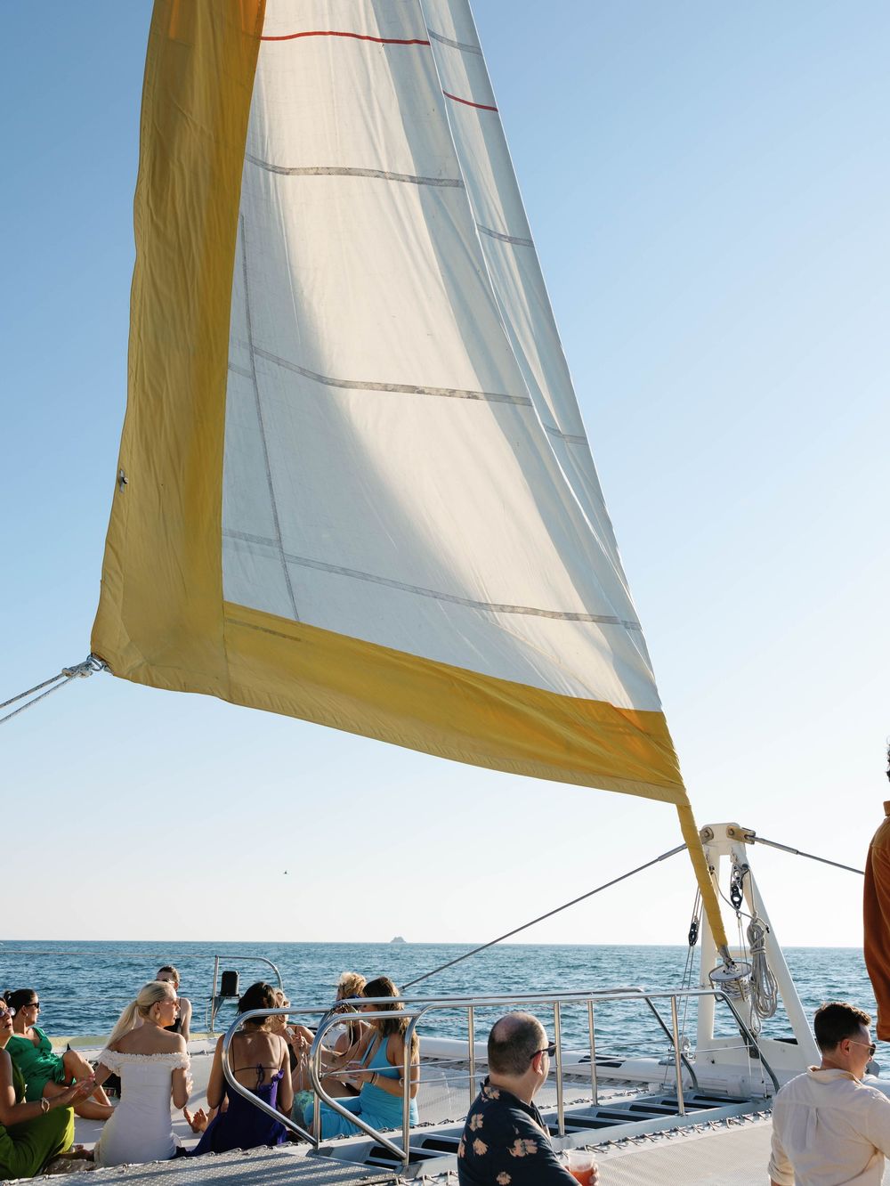White and yellow sail against blue sky with passengers visible on deck below.