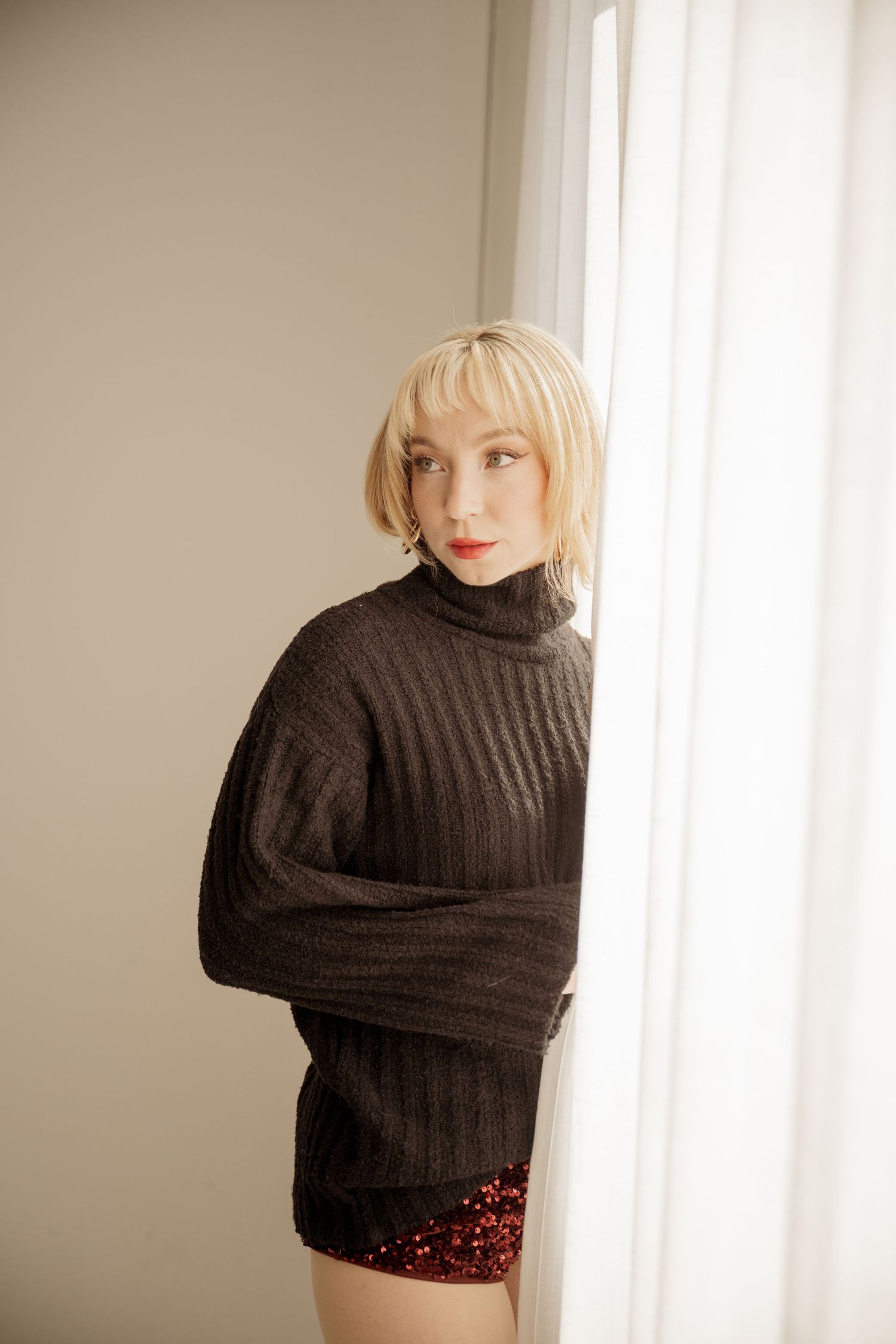 Person wearing cozy brown turtleneck sweater and red sequined shorts poses by bright window in soft lighting.