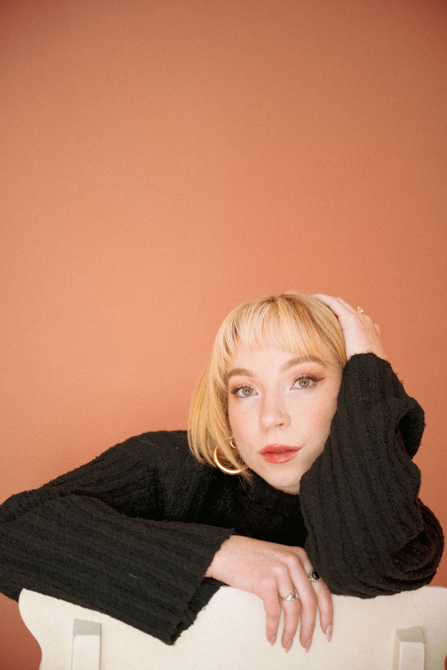 Portrait photo against peachy orange background showing someone in black sweater resting head on arm with soft expression.