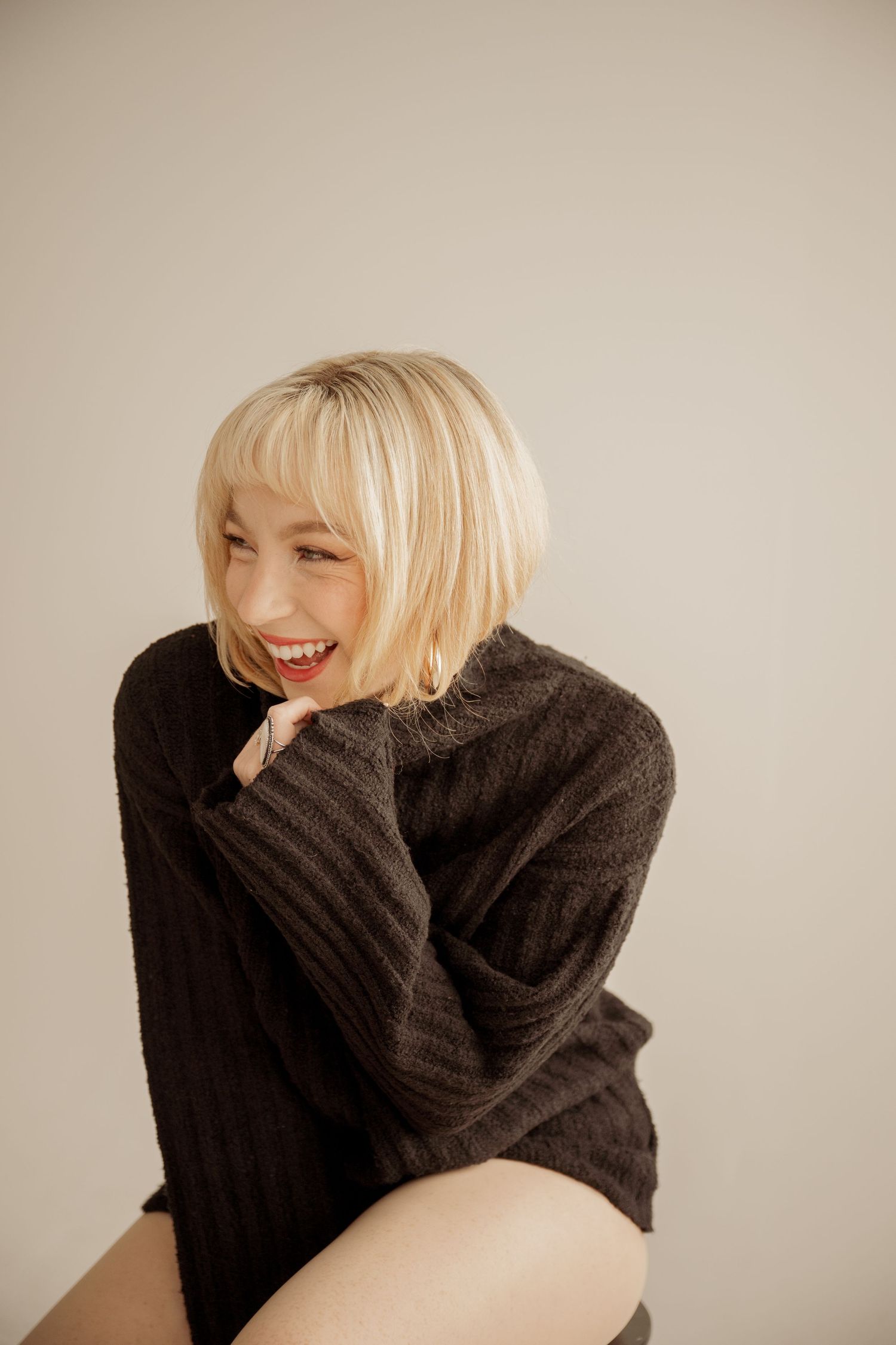 Happy person in black knit sweater laughs joyfully while sitting against neutral beige background.