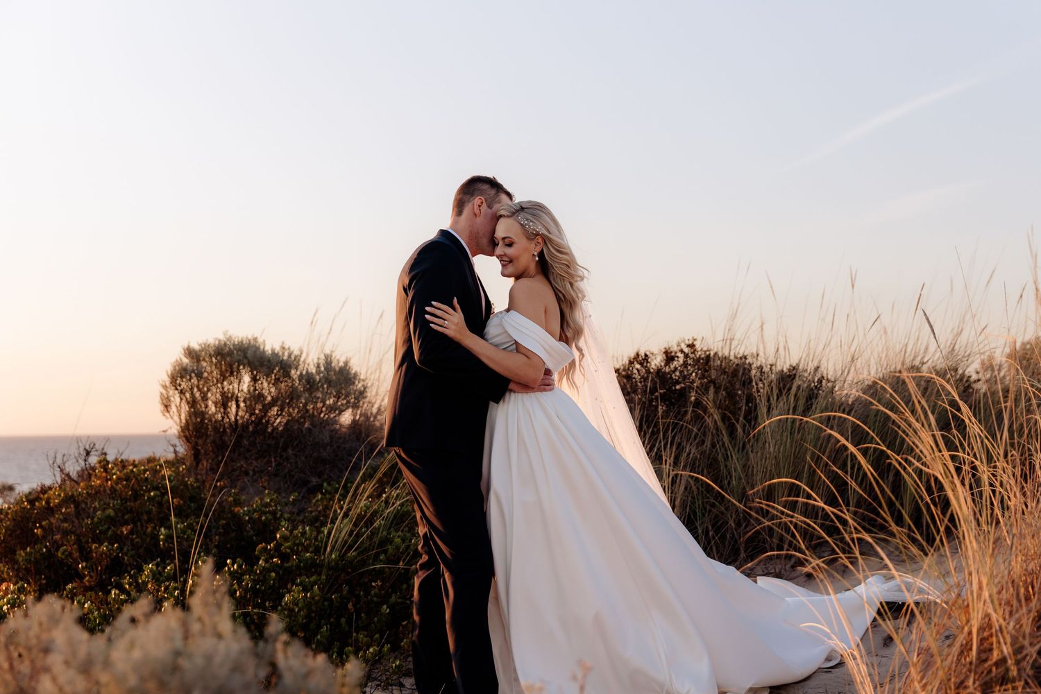 A couple embraces against a golden sunset on a hill with tall grass swaying in the breeze.