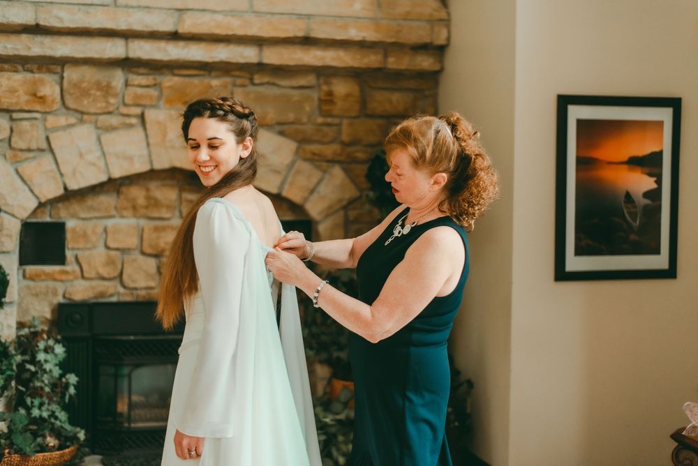 Mother of bride helps zip up bride's wedding dress in front of fireplace