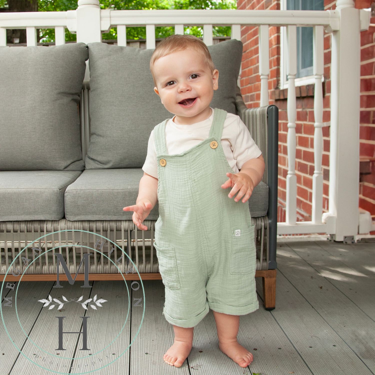 Toddler wearing mint green overalls stands smiling on a front porch with cushioned seating and brick walls.