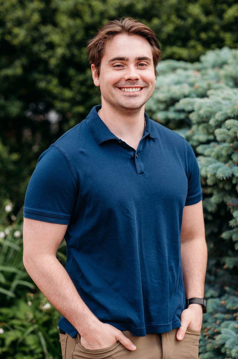 A person in a navy blue polo shirt poses outdoors with evergreen trees in the background.