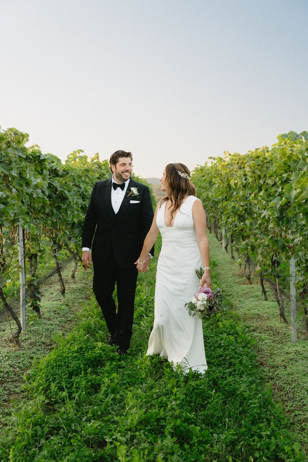 A couple walks hand in hand through lush vineyard rows in formal wedding attire.
