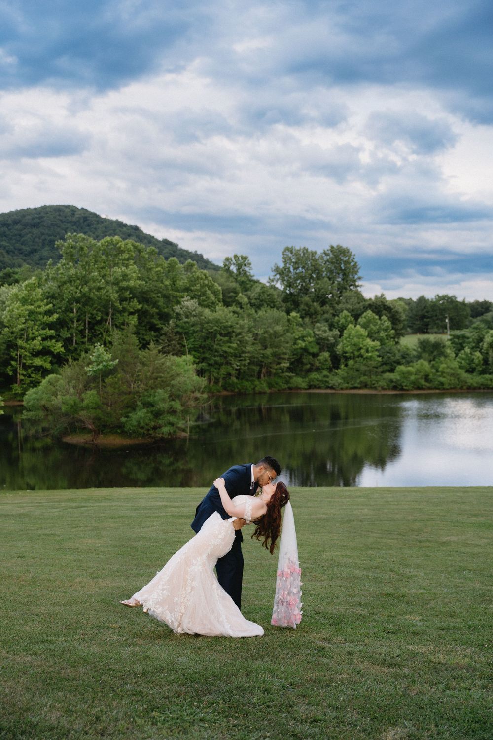 Romantic wedding couple shares a kiss by a serene lake with mountains and dramatic clouds in the background.