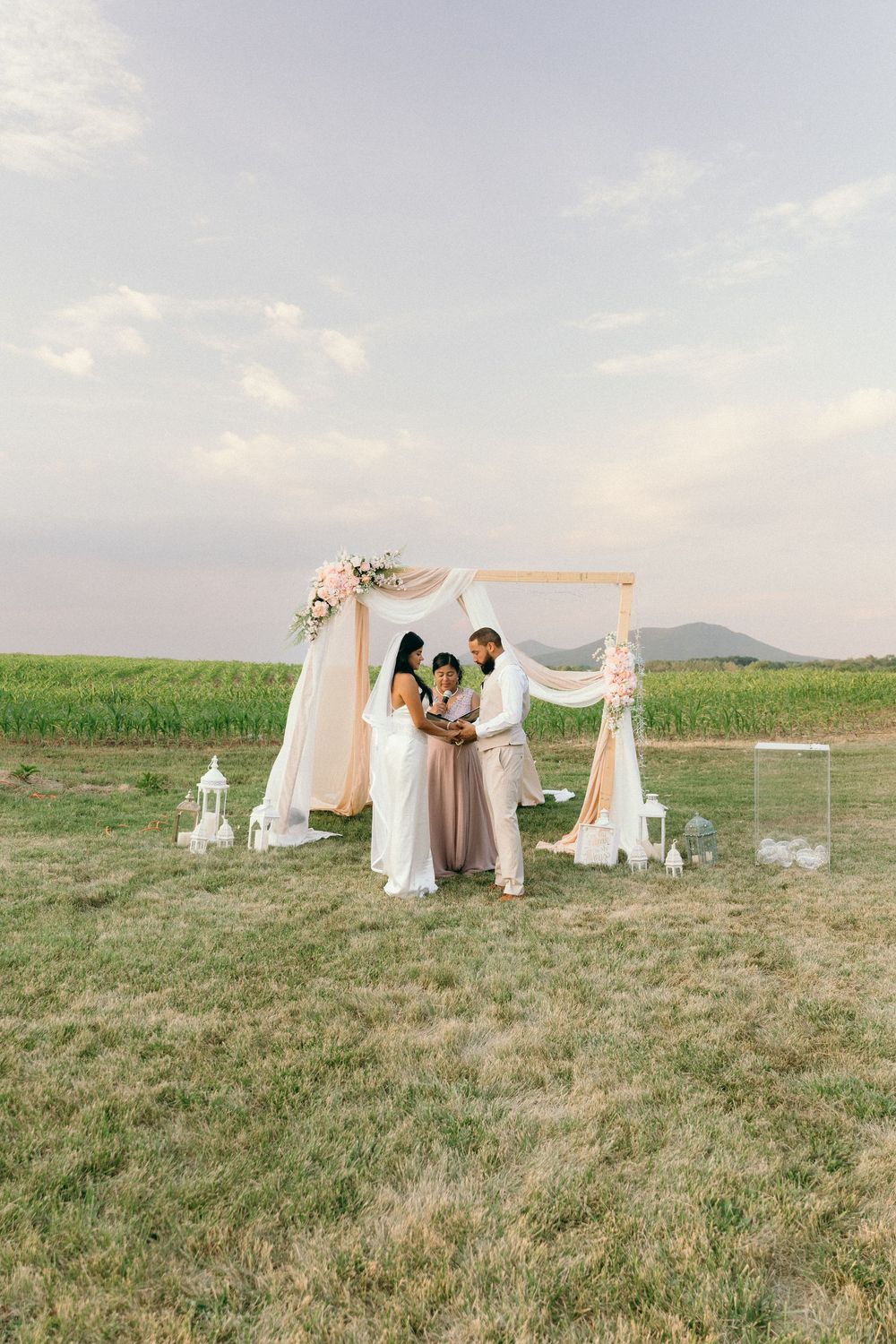 Wedding ceremony setup with white draped arbor in a rustic field at sunset.
