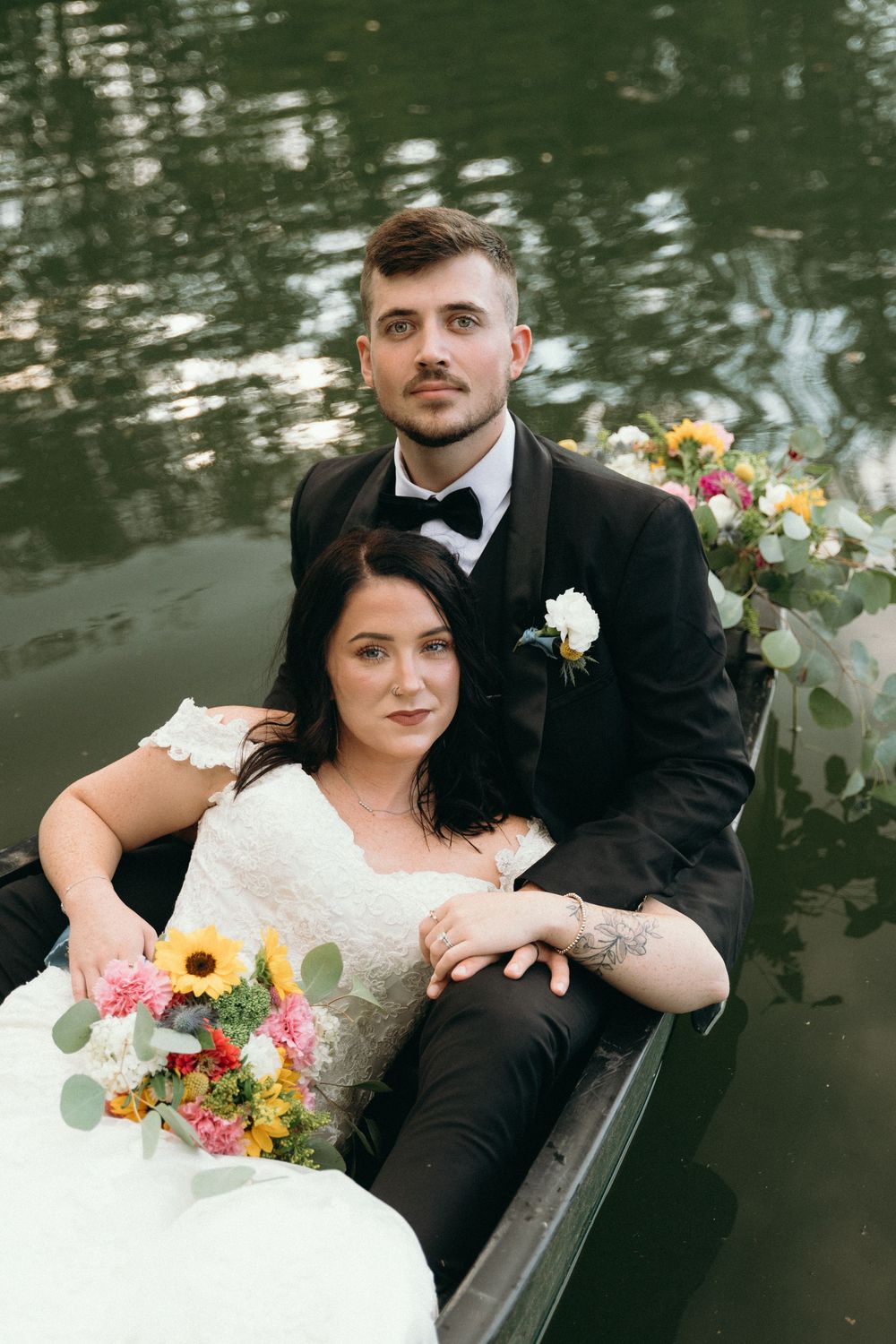 Wedding couple poses in wooden boat with colorful bouquet against reflective water.