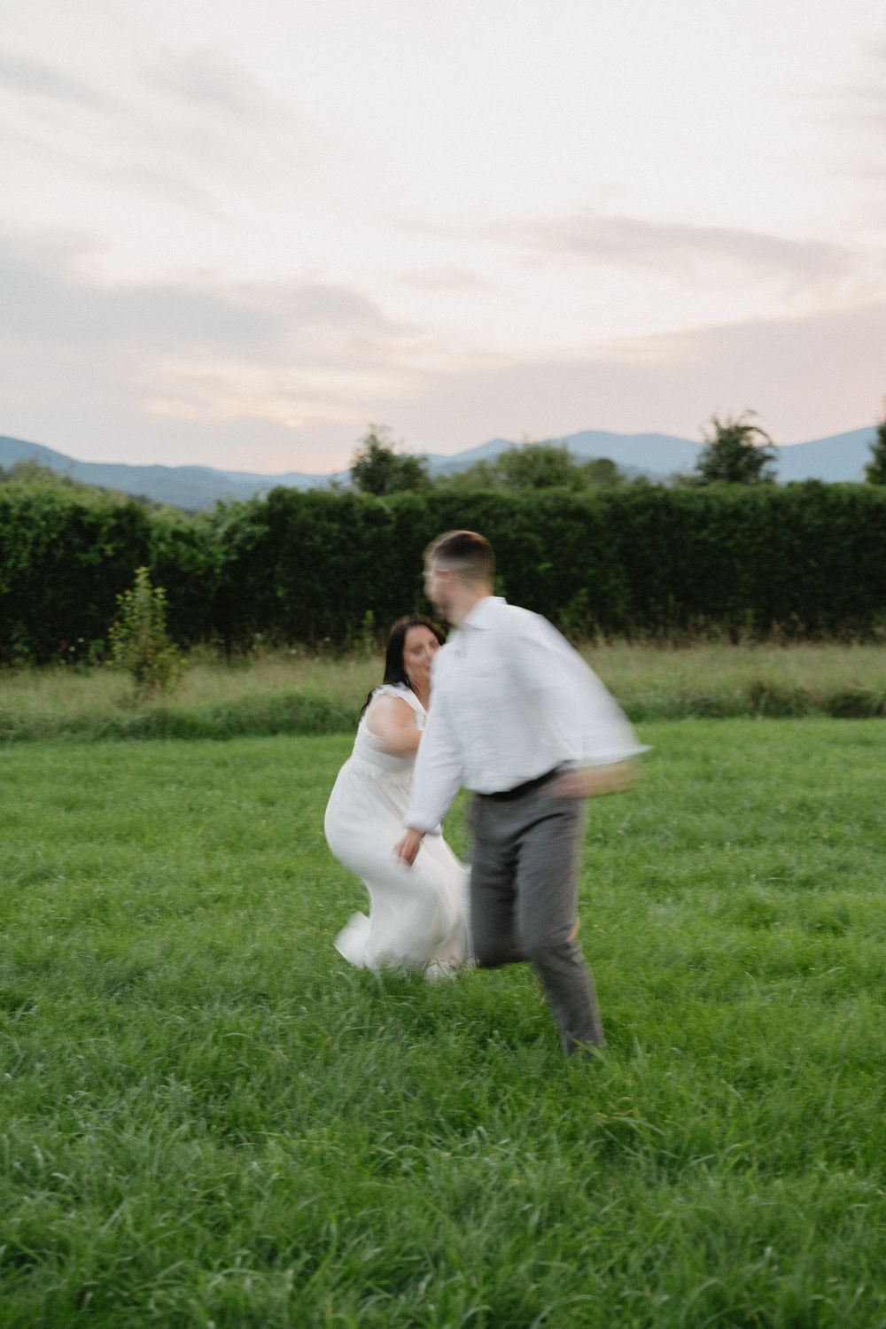 Playful wedding couple runs through green field with mountains in background at sunset.