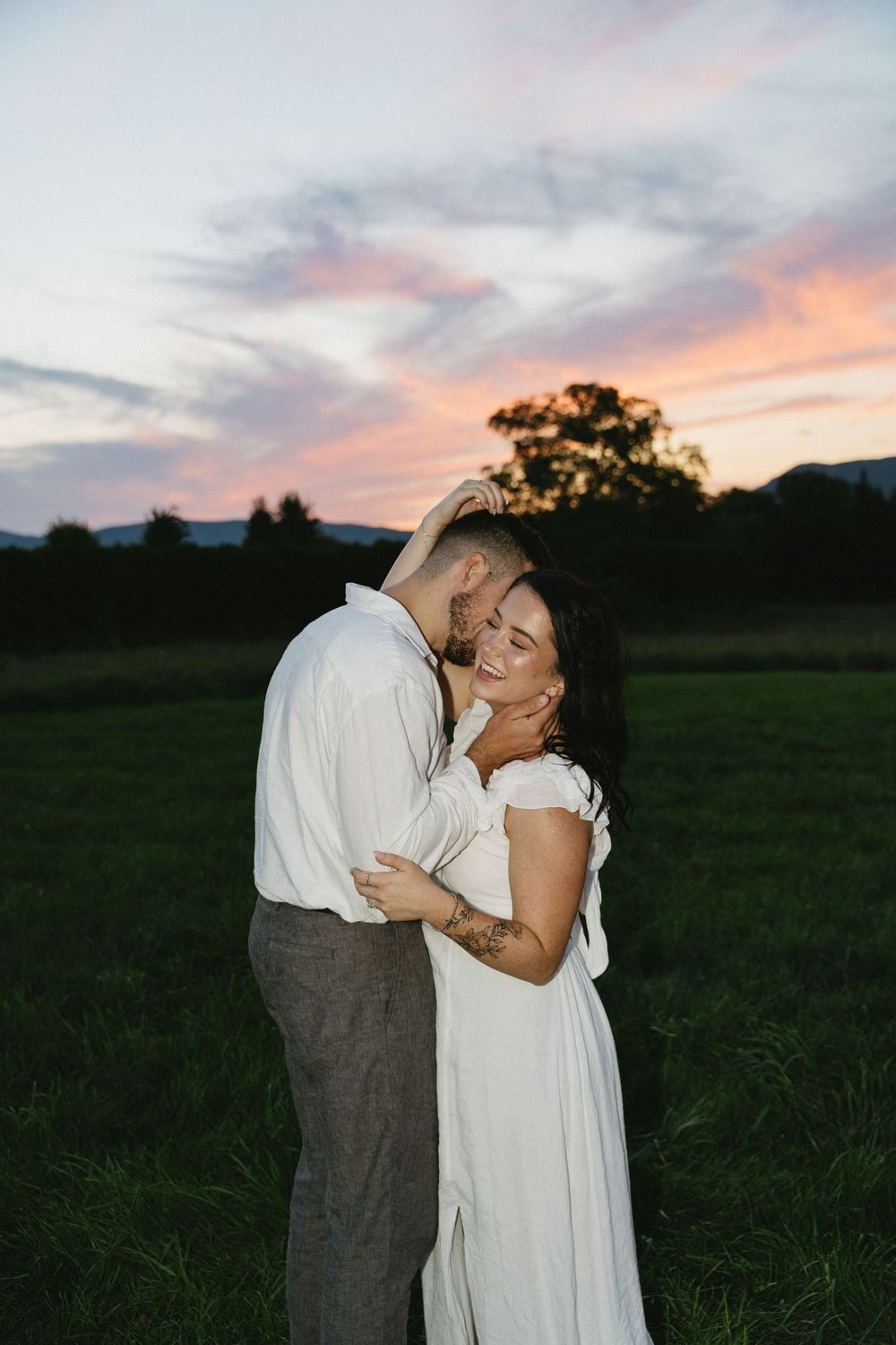 A couple embraces in a sunset-lit field with mountains and dramatic sky in the background.
