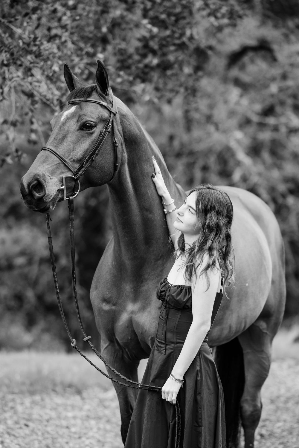 A beautiful black and white portrait of a woman in a dark dress standing beside an elegant horse in a natural setting.