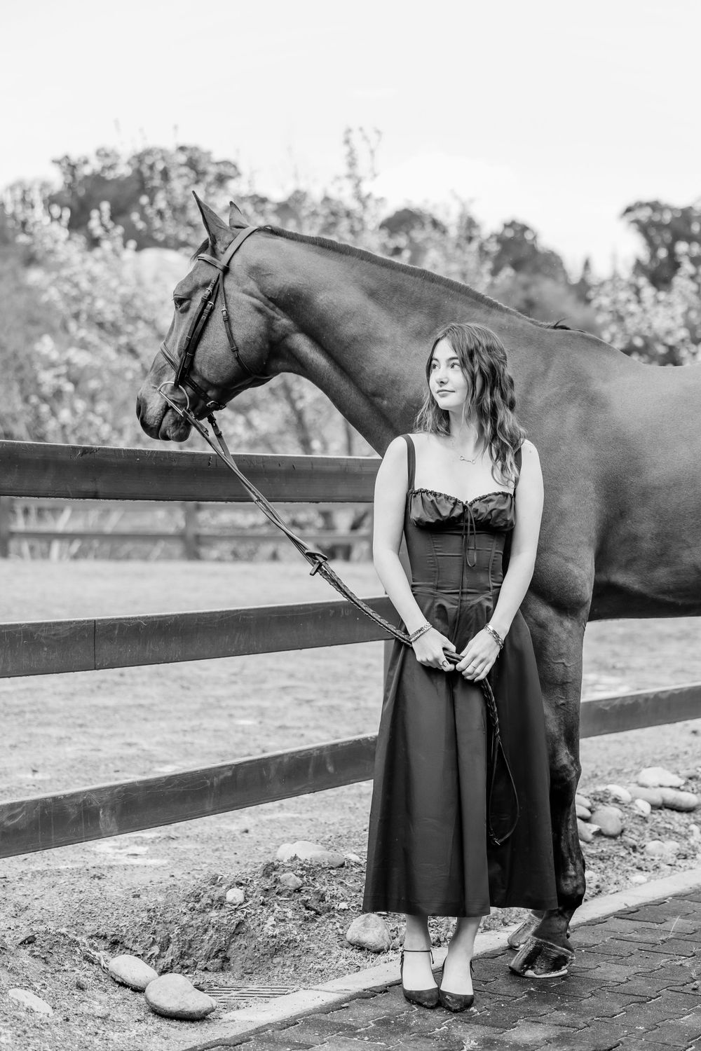 Black and white photo showing a figure in a long dress standing beside a dark horse near a wooden fence.