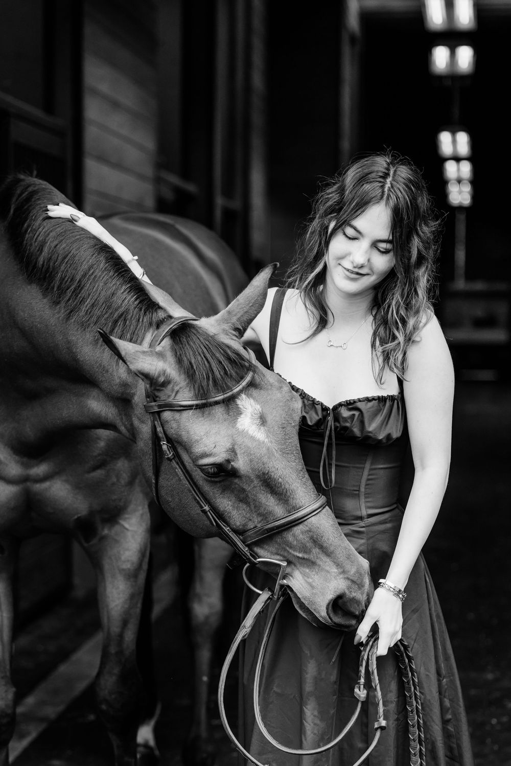 A tender black and white moment between a woman in elegant dress and a gentle horse outside a rustic stable.