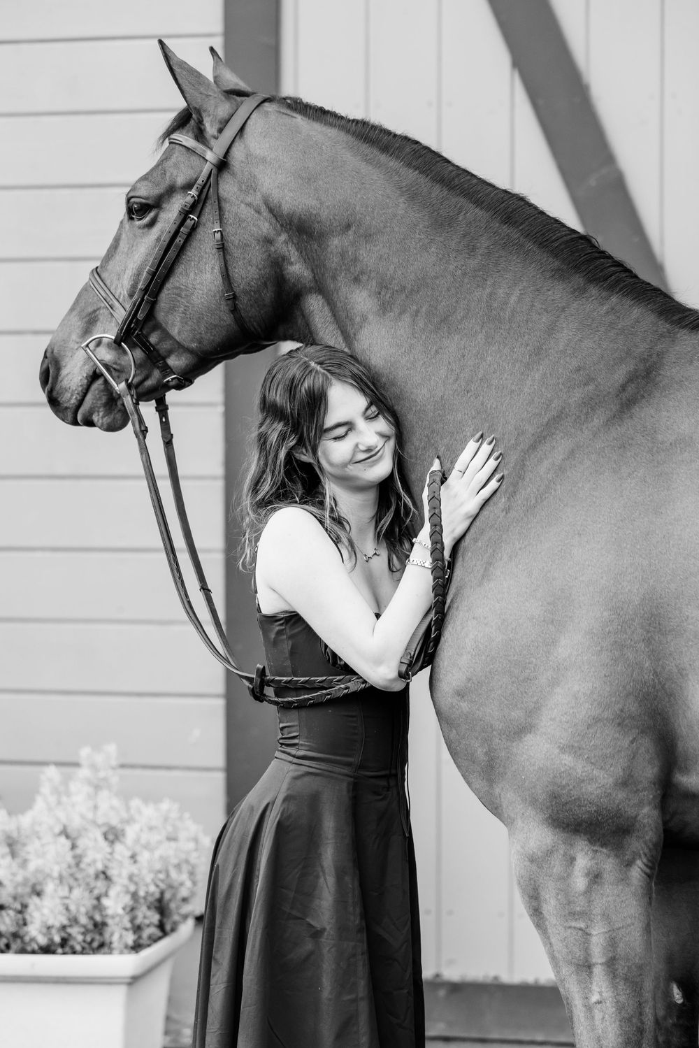 A woman in a black dress embraces a dark brown horse while standing next to a white building.