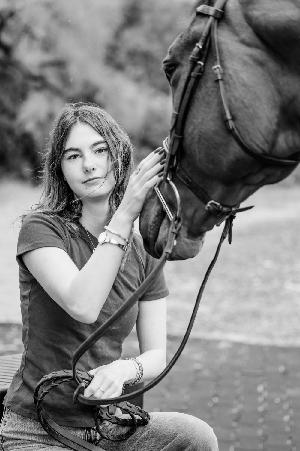 Black and white portrait of a person with long hair gently touching the face of a dark horse with bridle and reins.