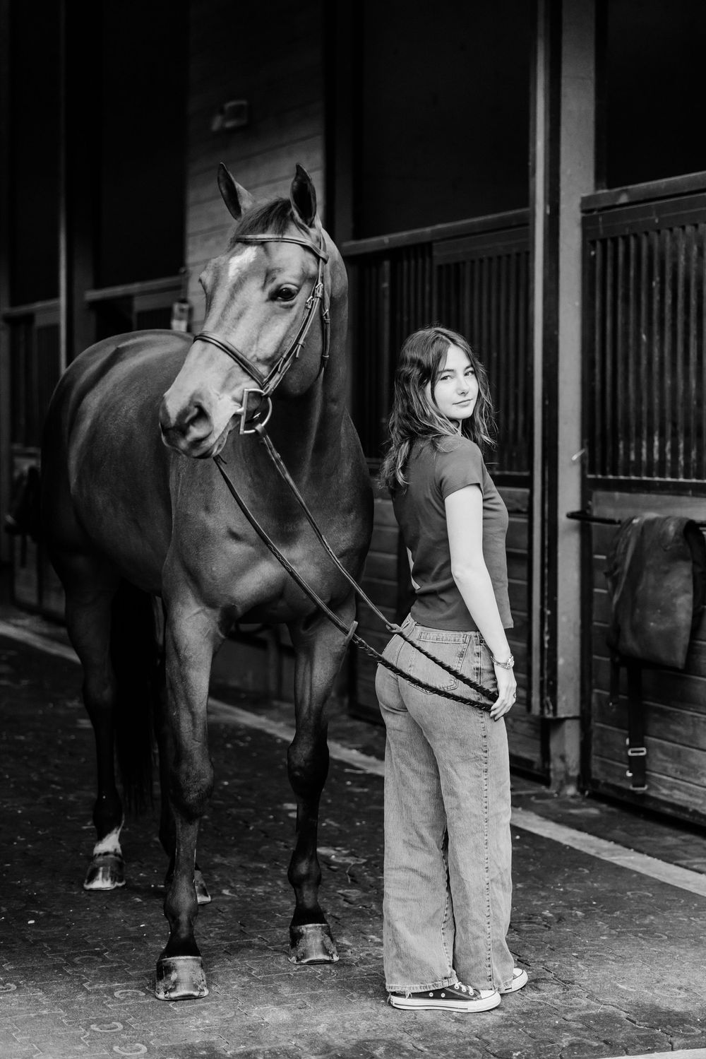 Black and white photo of someone standing beside a saddled horse in a stable hallway.