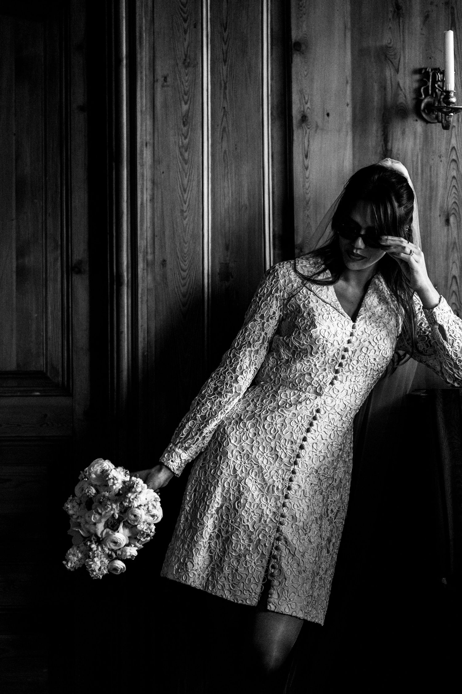 A moody black and white photo of someone in a lace dress holding a bouquet against wood paneled walls.