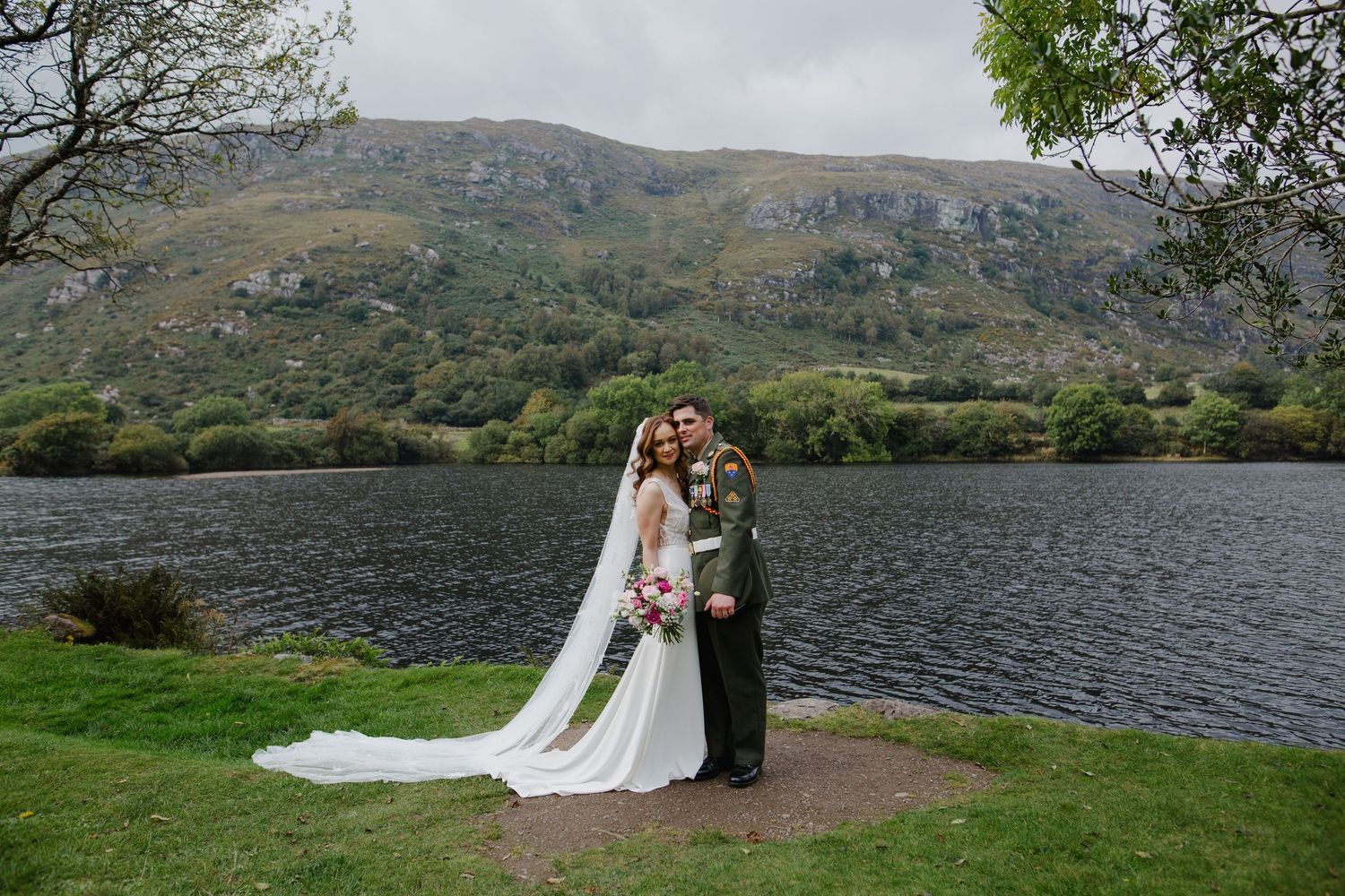 A couple stands together by a serene lake surrounded by rolling hills and mountains in a scenic outdoor setting.
