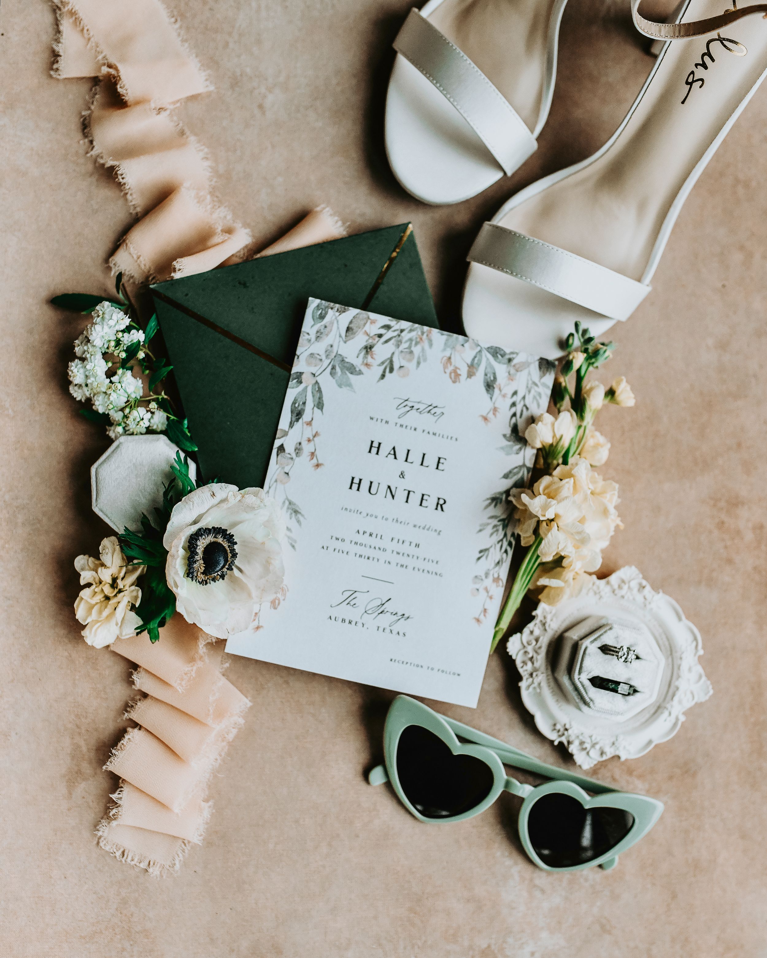 Flat lay of bride’s wedding details including engagement ring, wedding bands, invitation suite, and vintage ring box on a soft ivory fabric