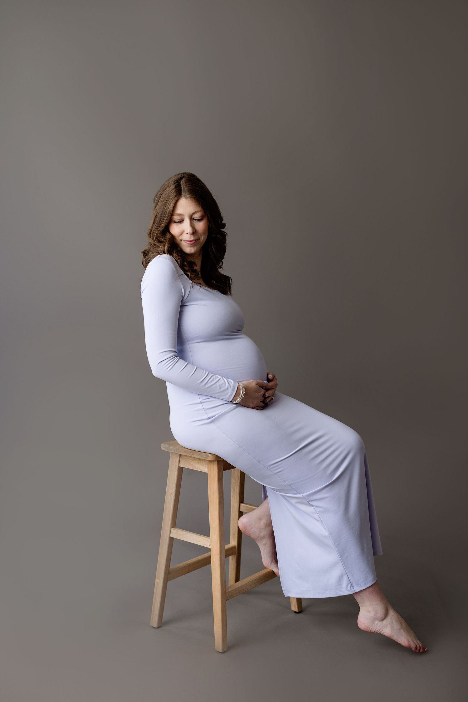 Person in long white dress sits gracefully on wooden stool against gray backdrop during maternity photoshoot.