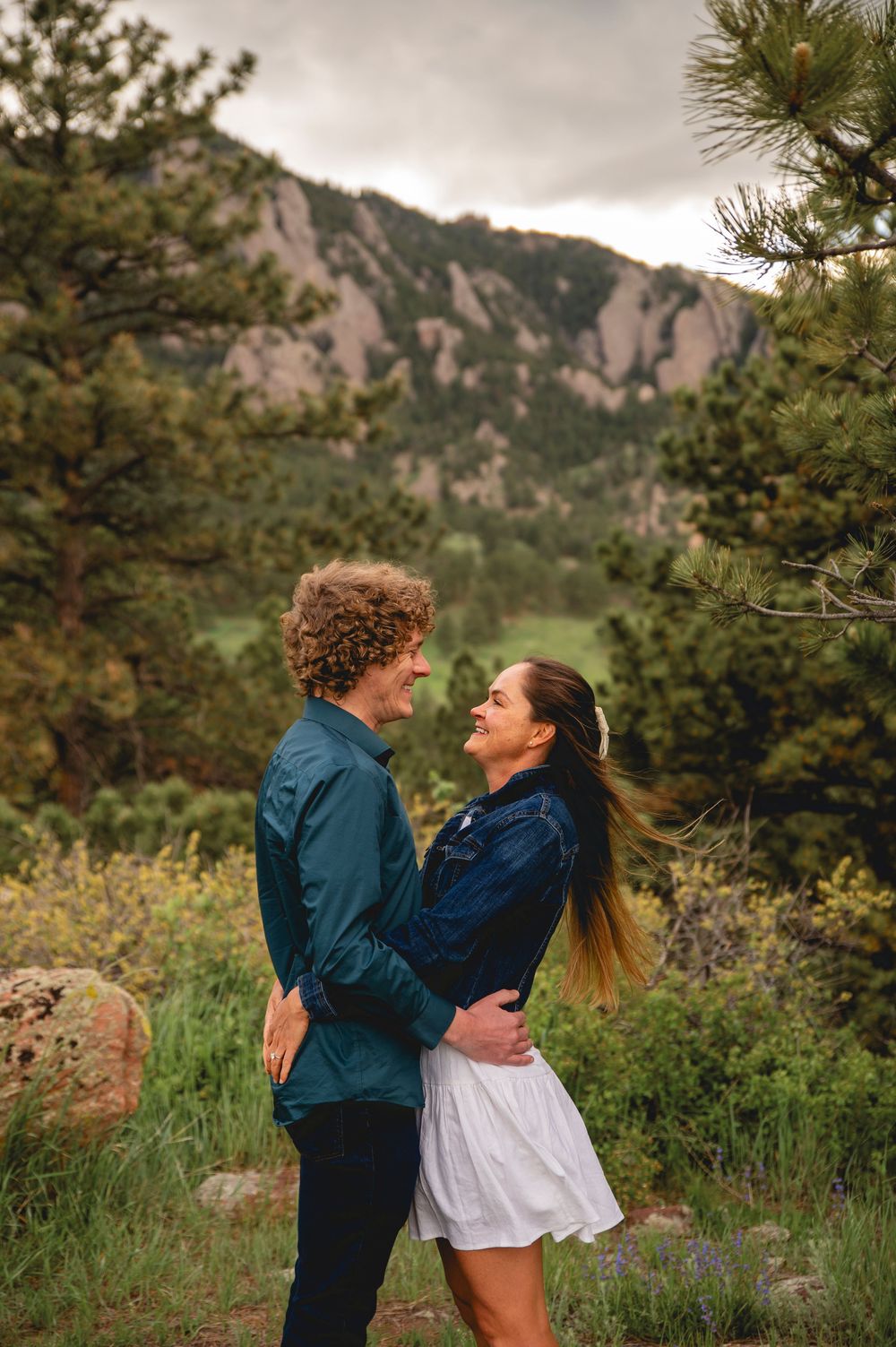 A couple embraces on a mountain trail with rugged cliffs and pine trees framing the scenic wilderness landscape.