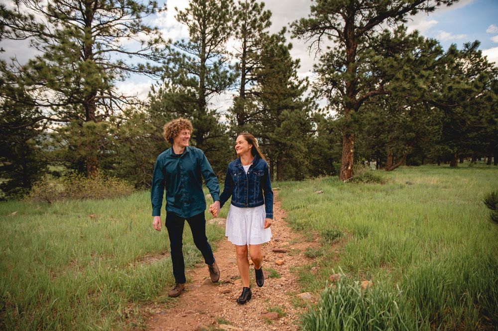 Two people walk together holding hands on a dirt trail through a grassy meadow surrounded by tall pine trees.