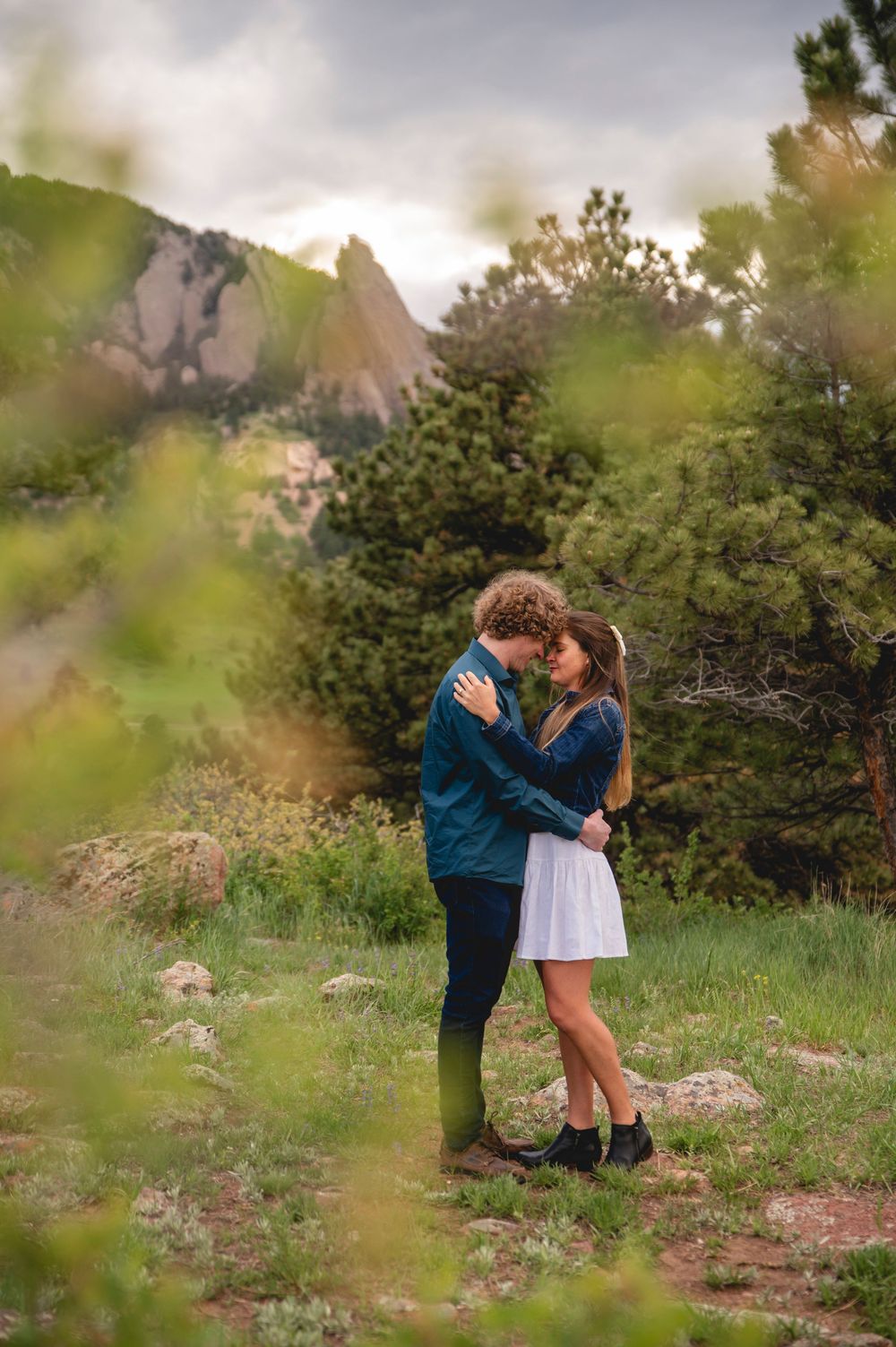 A couple embraces on a mountain trail surrounded by green foliage and rocky cliffs in the background.