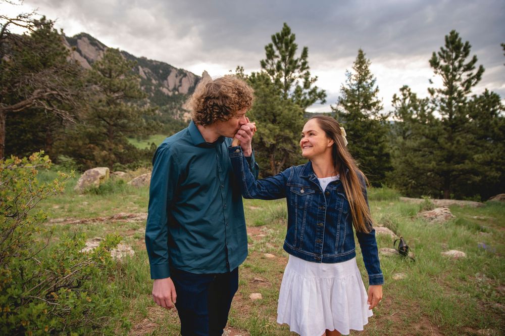 A couple shares a joyful moment while hiking in the mountains surrounded by pine trees and dramatic cloudy skies.