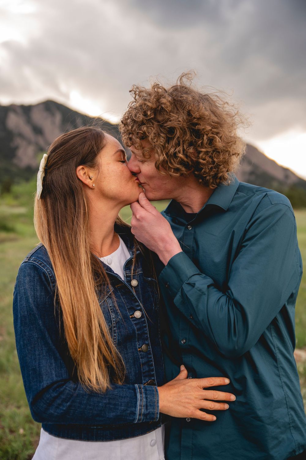 A couple sharing a tender kiss in a field with mountain landscape and cloudy skies in the background.