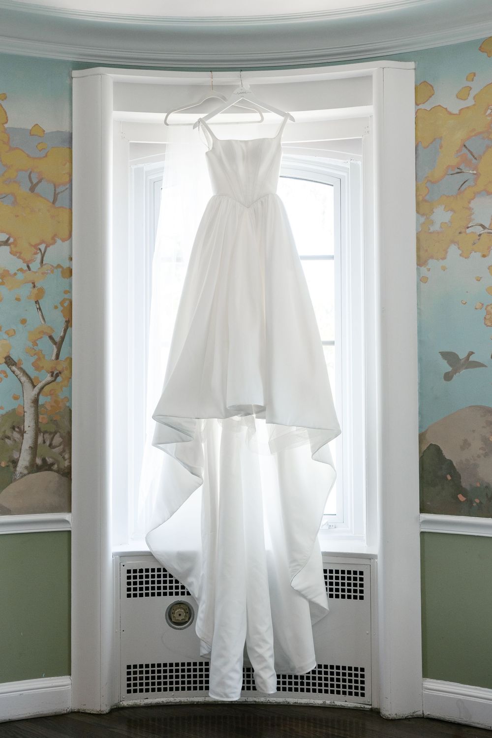 Classic white wedding dress hanging in bay window with elegant wallpapered walls.