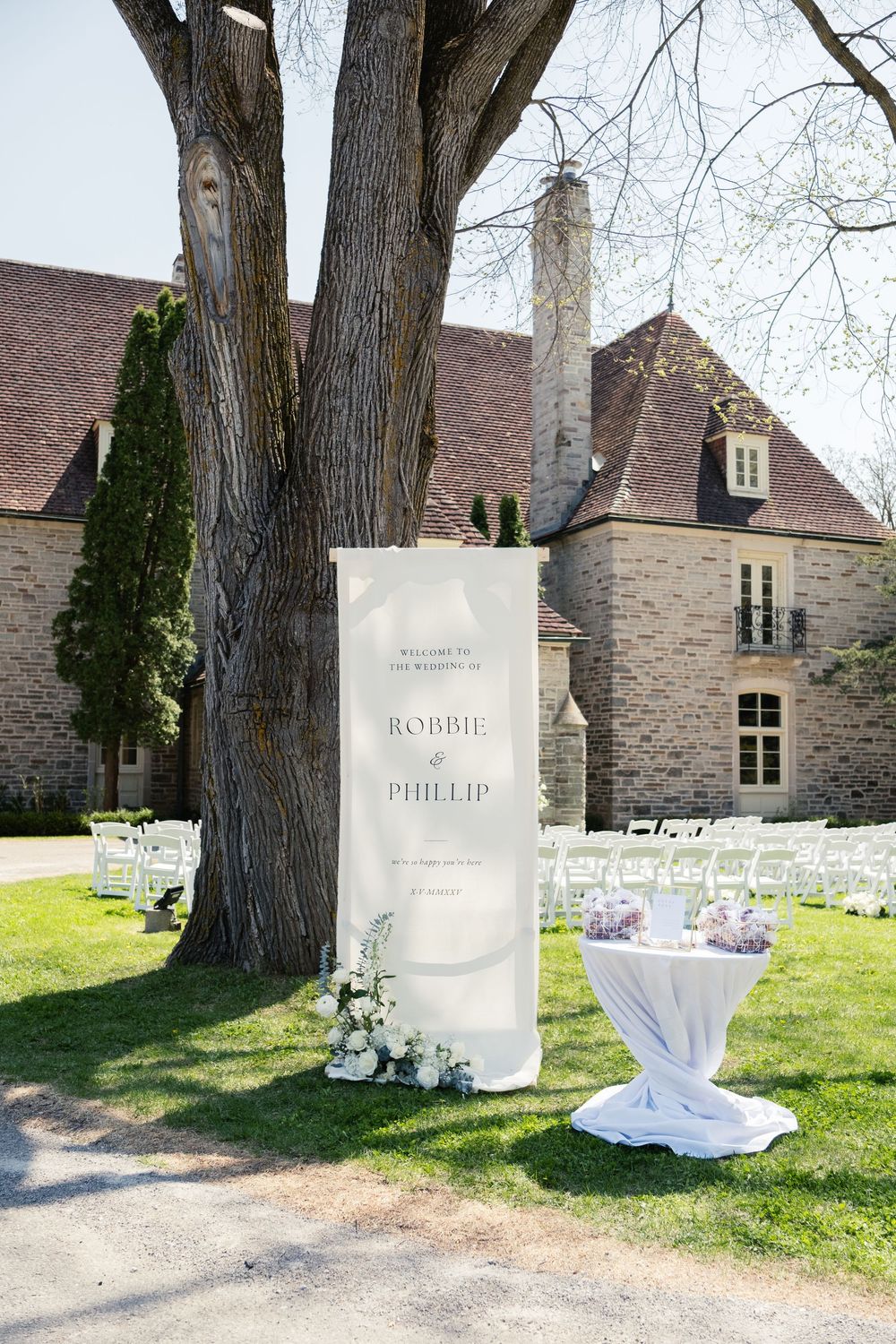 A wedding welcome sign and cocktail table arranged outdoors near a large tree and stone building.