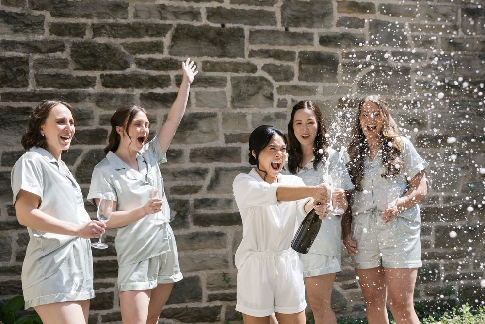 Group celebrates with champagne spray outside stone building in matching white outfits.