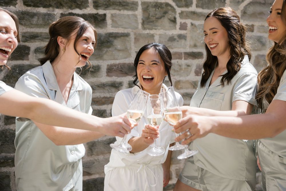 Wedding party in white robes toasts with champagne glasses against stone wall.