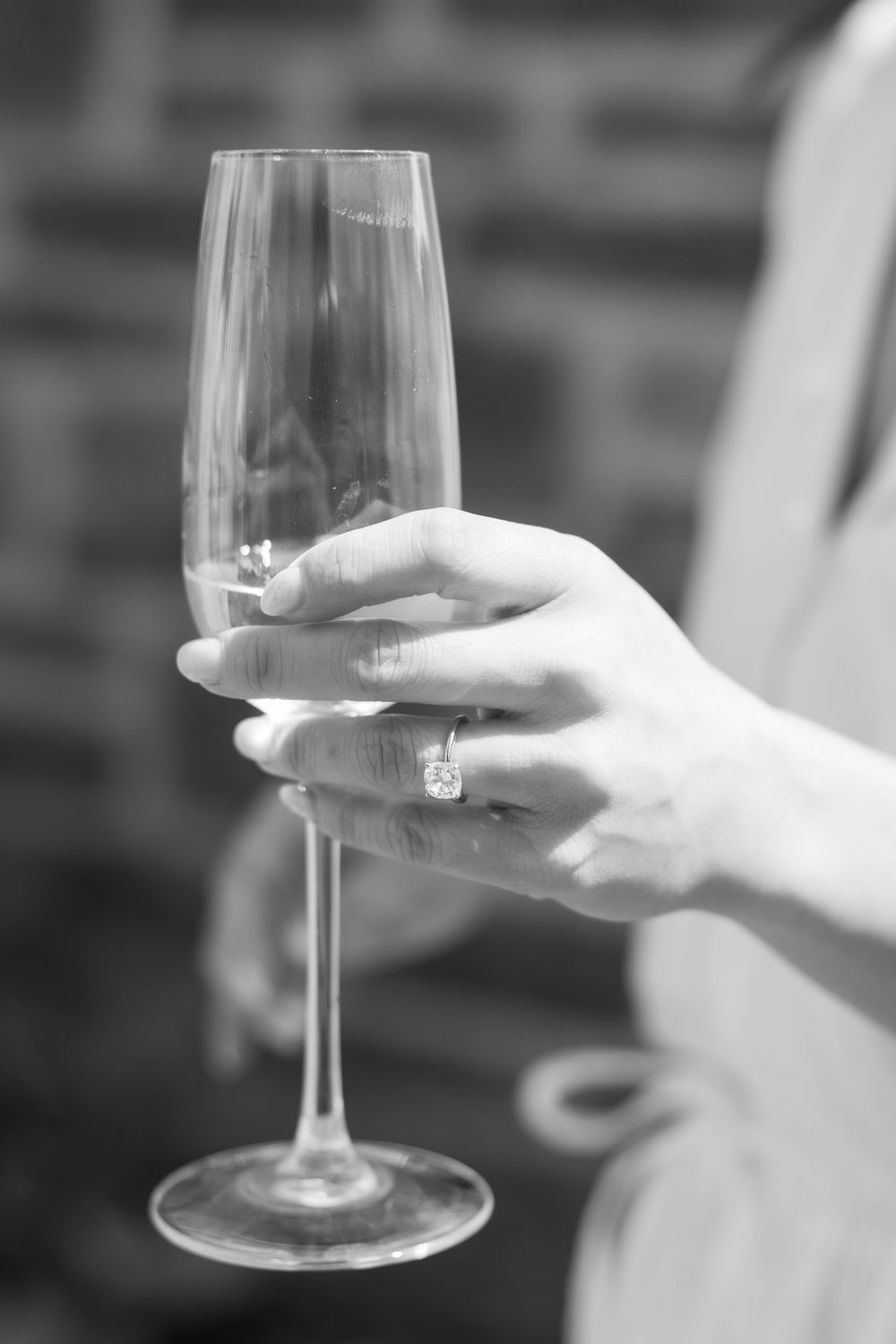 Close-up of champagne glass held by hand with engagement ring in black and white.