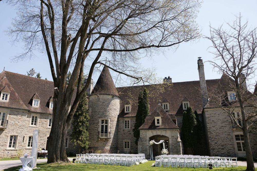Stone mansion wedding venue with tall trees and white chairs set up for an outdoor ceremony.