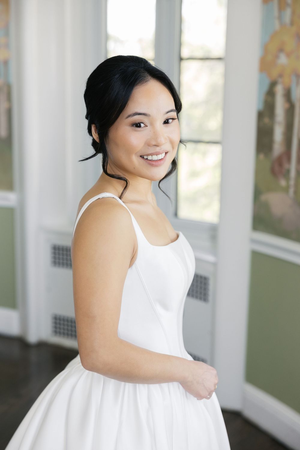 Woman in white wedding dress stands by window in bright interior.